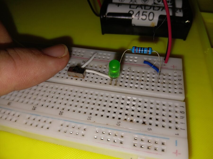 A hand pressing a wire into a breadboard beside a lit green LED and a battery pack, from testing the curriculum on the bench